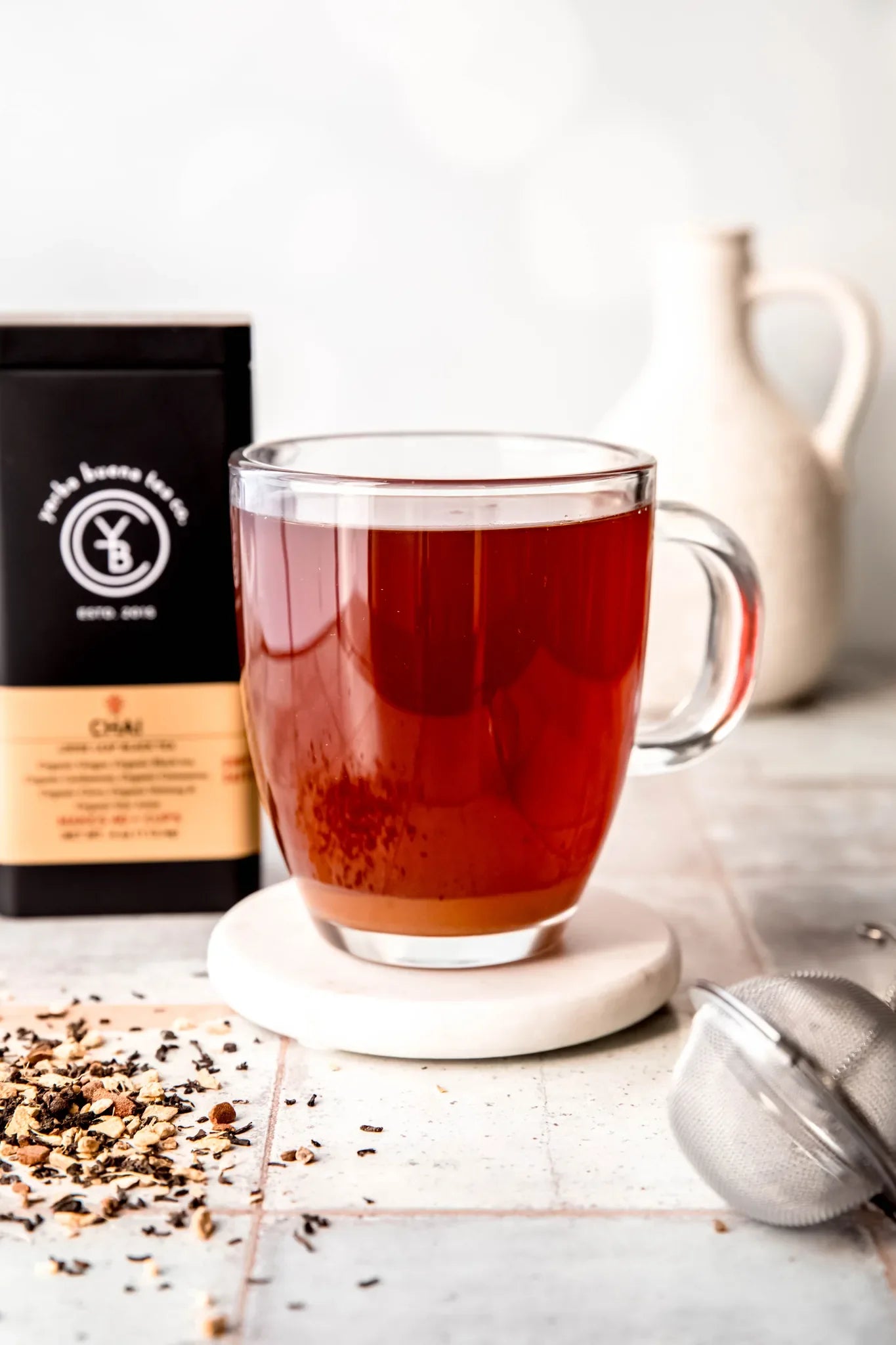 Steaming cup of deep red Chai tea on a white coaster, with loose leaf tea and branded pouch displayed on a light surface, by Yerba Buena Tea Company.