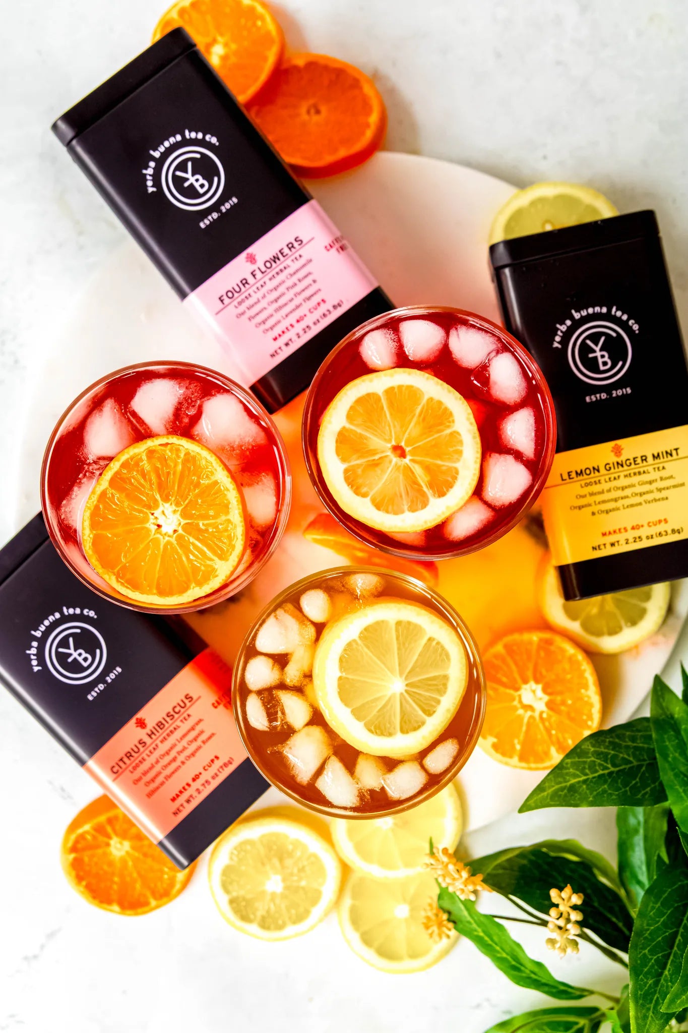 Three iced tea glasses with orange and lemon slices and ice cubes on a marble surface, surrounded by tea packages including Four Flowers and Lemon Ginger Mint, fresh citrus slices, and green leaves, from Yerba Buena Tea Company.