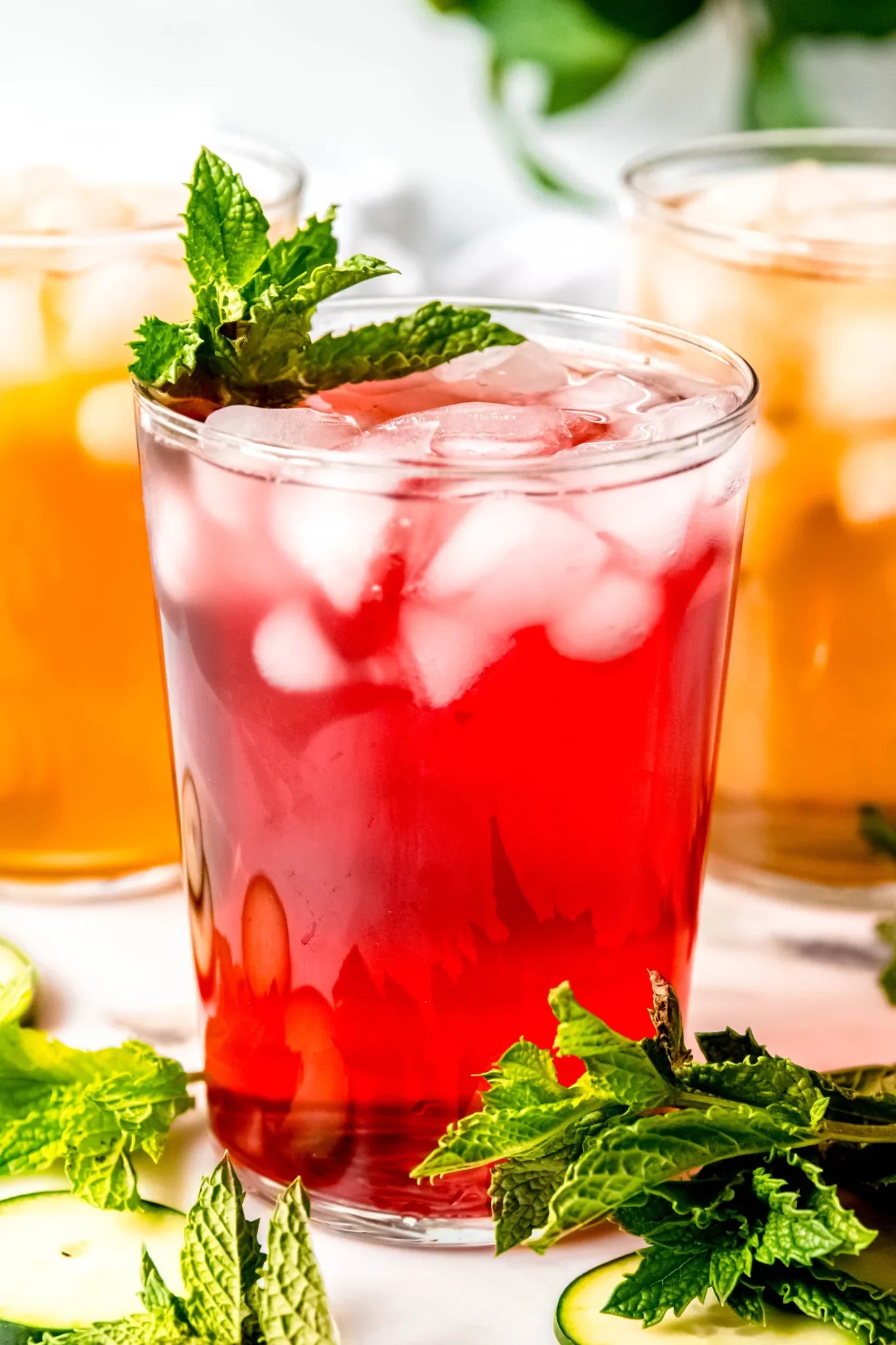A tall glass of bright red iced hibiscus tea garnished with fresh mint leaves and ice cubes, surrounded by loose mint sprigs on a sunlit counter, from Yerba Buena Tea Company.