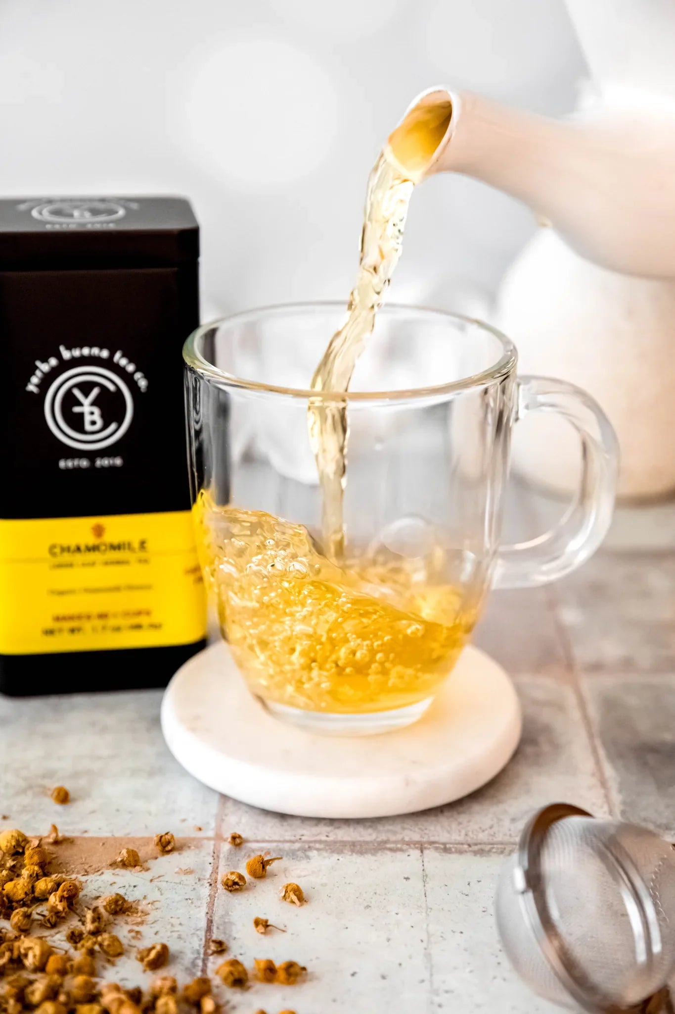 Chamomile herbal tea steaming into a glass mug from a ceramic pouch, with dried chamomile flowers scattered on a light tile surface, by Yerba Buena Tea Company.
