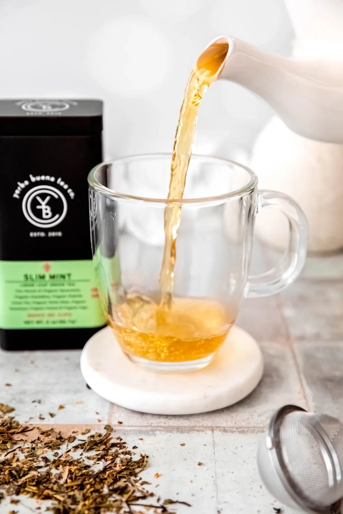 Slim Mint yerba mate tea being steeped in a clear glass mug on a white coaster, with loose leaf herbs scattered on a light concrete surface, by Yerba Buena Tea Company.