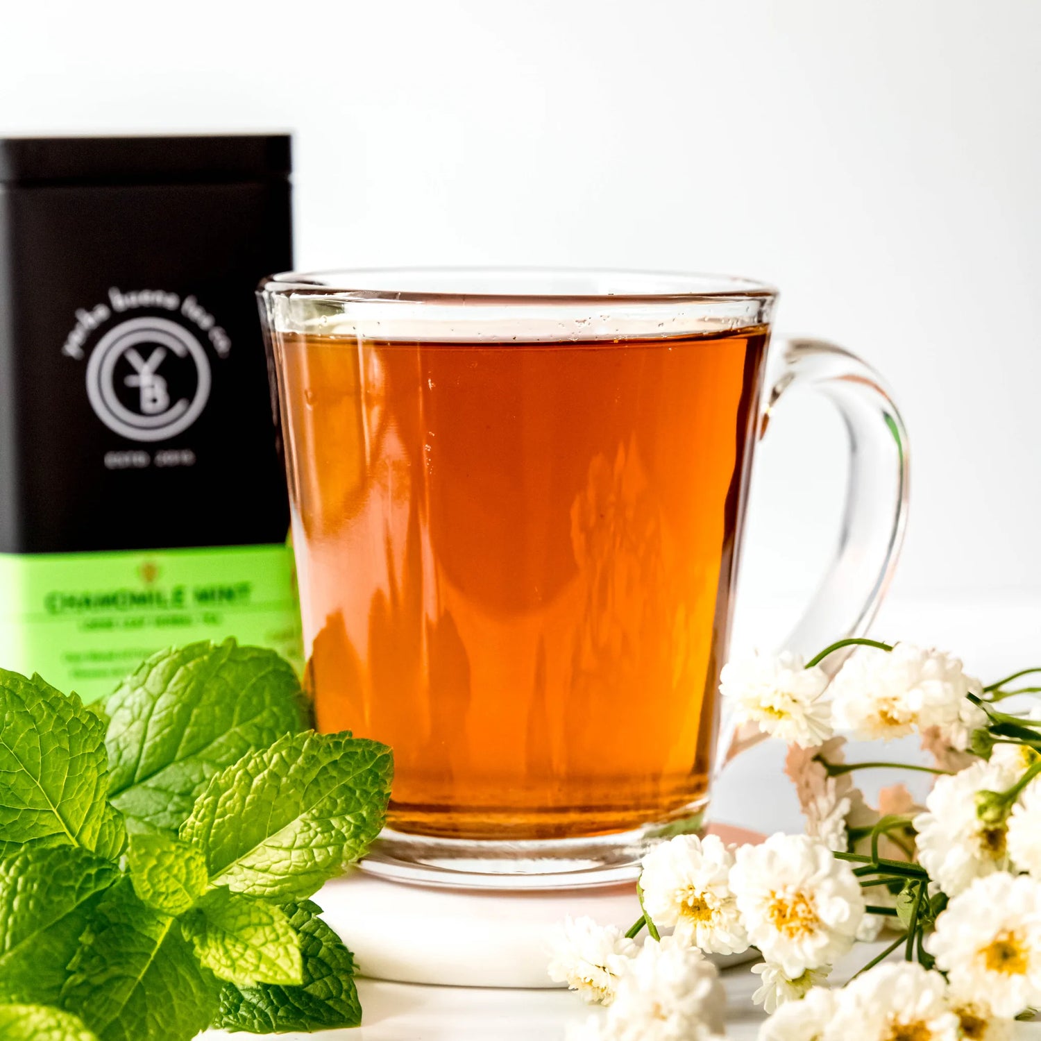 Brewed chamomile mint tea in clear glass mug with fresh mint leaves and white flowers nearby, by Yerba Buena Tea Company.