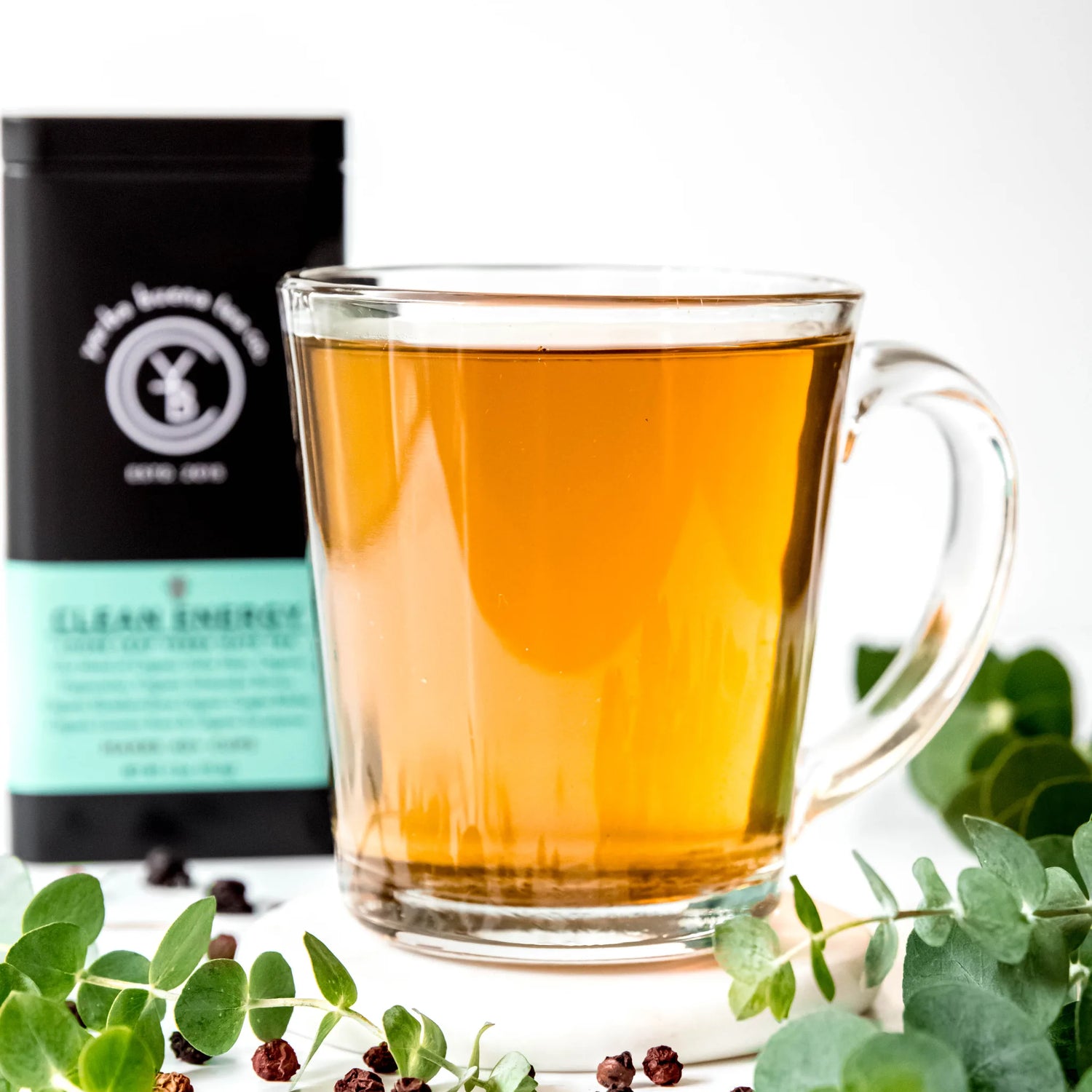 Brewed golden tea in a clear glass mug surrounded by fresh green leaves and packaging, by Yerba Buena Tea Company.