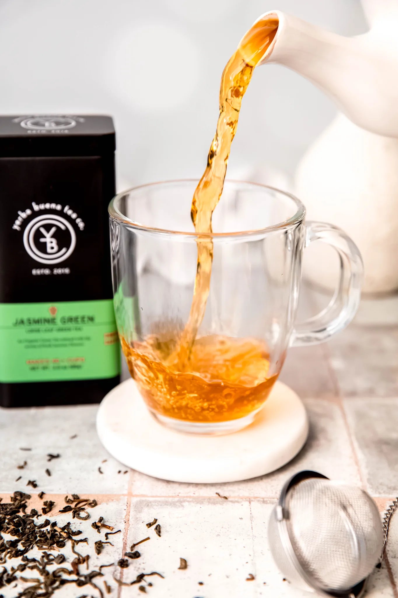 Jasmine Green tea steeping in a clear glass mug as golden liquid pours from a white teapot, with loose leaf tea scattered on marble surface below, by Yerba Buena Tea Company.