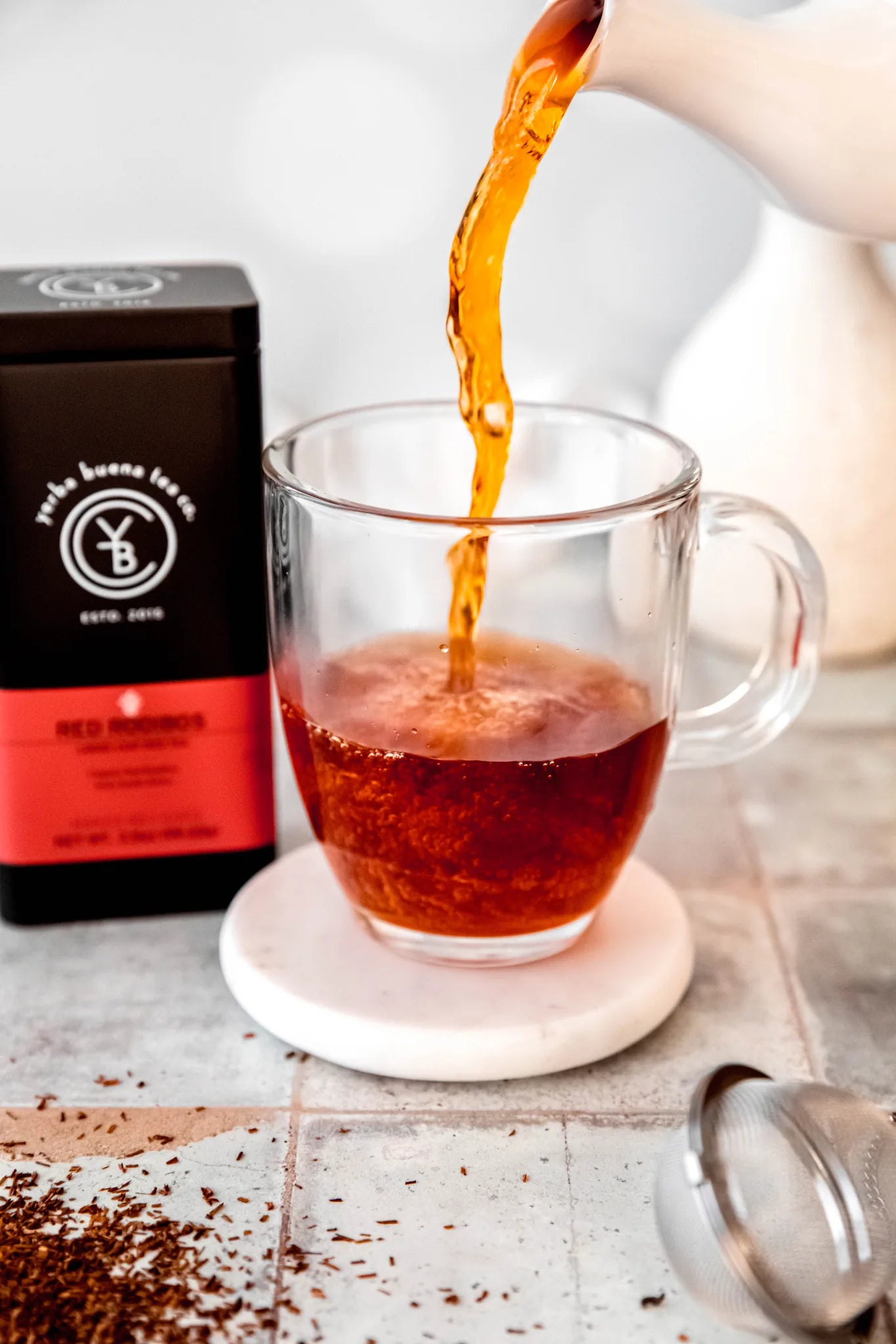 Rooibos Teas collection: amber liquid pours into a clear mug on a white coaster, with a branded black and red tea pouch and loose leaves on a light concrete surface, by Yerba Buena Tea Company.