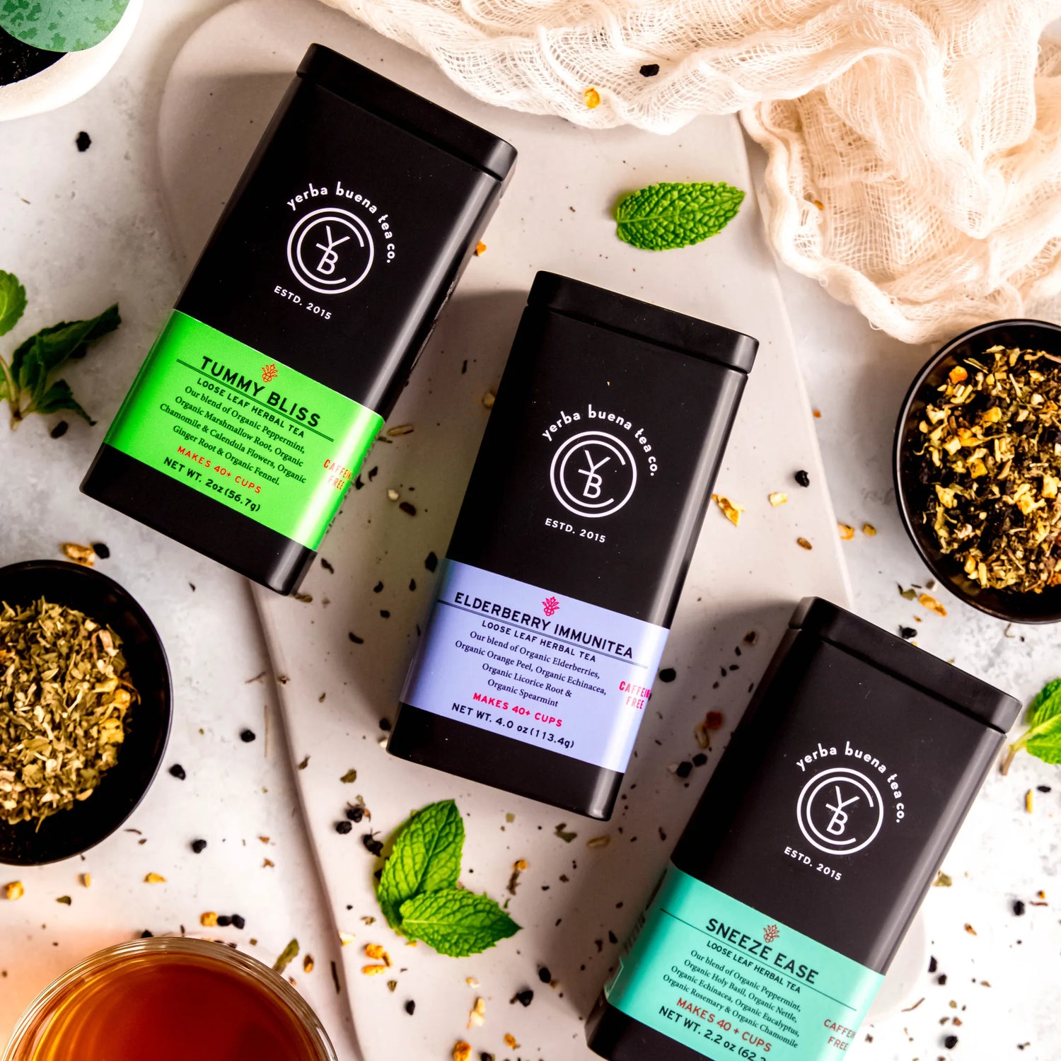 The Wellness Feel Better Box Set gift collection featuring three loose leaf herbal teas: Tummy Bliss, Elderberry Immunitea, and Sneeze Ease in branded canisters, arranged with fresh mint leaves and dried botanicals, by Yerba Buena Tea Company.