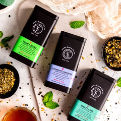 The Wellness Feel Better Box Set gift collection featuring three loose leaf herbal teas: Tummy Bliss, Elderberry Immunitea, and Sneeze Ease in branded canisters, arranged with fresh mint leaves and dried botanicals, by Yerba Buena Tea Company.